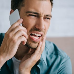 young man talking by smartphone and looking away while suffering from tooth pain at home